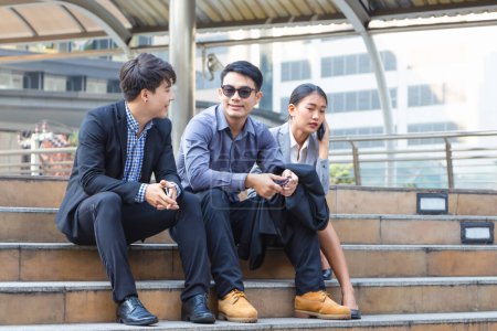 Foto de Jóvenes empresarios sentados en las escaleras con amigos, gente de negocios reuniéndose y compartiendo ideas, equipo de negocios de jóvenes asiáticos discutiendo por la mañana - Imagen libre de derechos