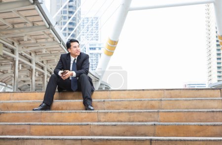Téléchargez les photos : Asiatique homme d'affaires assis sur les escaliers en dehors du bureau brouillé fond de la ville - en image libre de droit