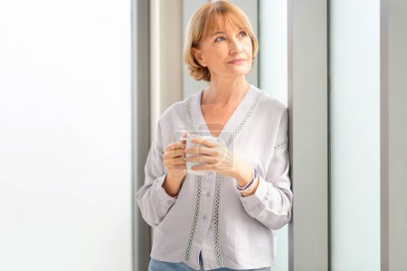 Téléchargez les photos : Femme relaxante debout près de la fenêtre avec des tasses de café, femme à l'intérieur de la nouvelle maison pendant une pause café - en image libre de droit