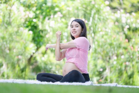 Téléchargez les photos : Asiatique femme enceinte méditant tout en étant assis dans le parc, femme faisant des exercices d'étirement à l'extérieur, Méditer sur la maternité - en image libre de droit