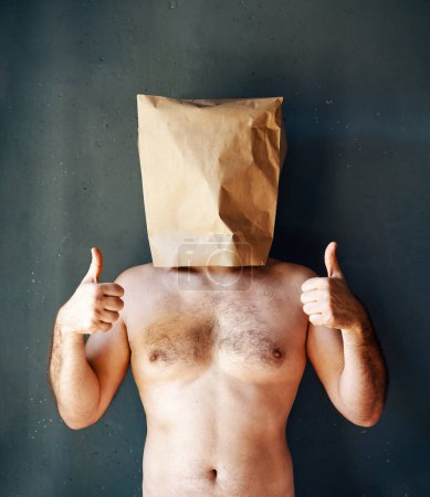 Foto de Hombre sin camisa con una bolsa de papel en la cabeza mostrando los pulgares hacia arriba sobre el fondo gris. Concepto de signo de aprobación - Imagen libre de derechos