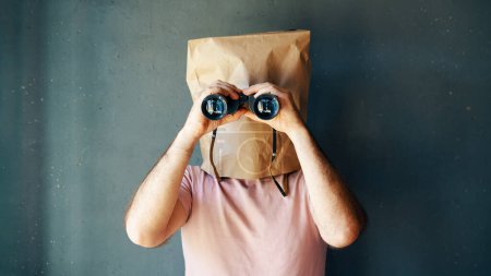 Man with a paper bag on head looking through binoculars to camera over gray background