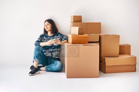 Smiling young woman enjoy her new home sitting on floor surrounded by cardboard boxes on moving day