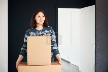 Young smiling woman moving into new apartment carrying cardboard boxes. Relocation concept