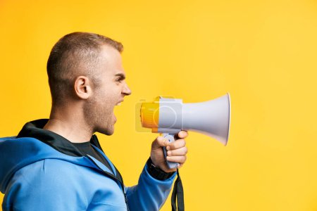 Young angry man shouting and screaming loud holding megaphone over yellow background