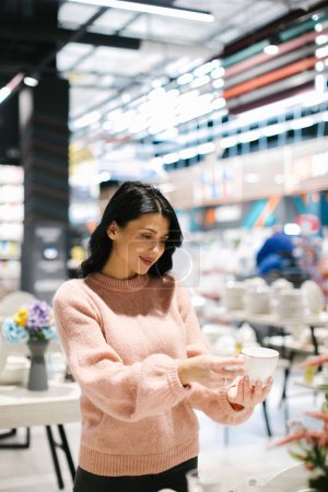 Téléchargez les photos : Une jeune femme brune en pull rose achète de la vaisselle dans un magasin. - en image libre de droit