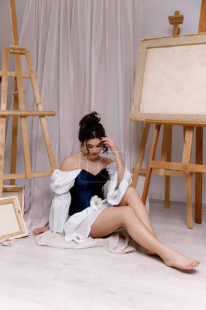 Foto de Una chica con el pelo largo y rizado con maquillaje en un vestido blanco con un corsé está sentada cerca de caballetes en un estudio de arte. - Imagen libre de derechos