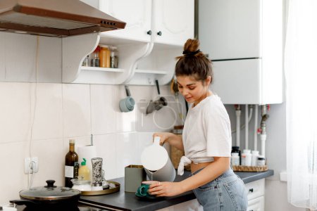 Photo for A brunette girl with hair styled in a bun is making tea in the kitchen in the morning. A girl in a white T-shirt and jeans is making tea in a white kitchen. - Royalty Free Image