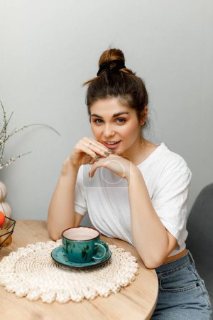 Téléchargez les photos : Une fille brune coiffée dans un chignon est assise dans la cuisine et boit du thé. Une fille en t-shirt blanc est assise à la table avec une tasse de thé vert. - en image libre de droit