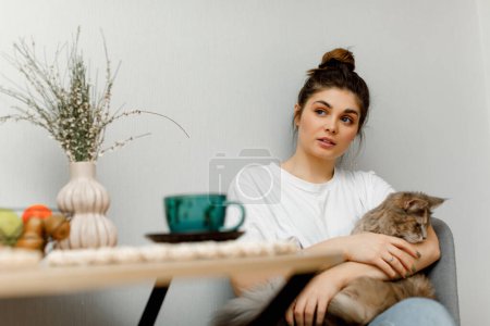 Téléchargez les photos : Une fille brune coiffée dans un chignon est assise dans la cuisine avec son chat et boit du thé. Une fille en t-shirt blanc est assise à la table avec une tasse de thé vert. - en image libre de droit