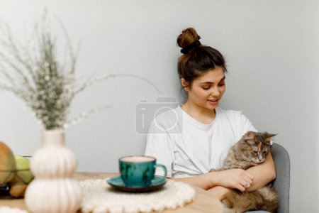 Photo for A brunette girl with hair styled in a bun is sitting in the kitchen with her cat and drinking tea. A girl in a white T-shirt is sitting at the table with a cup of green tea. - Royalty Free Image