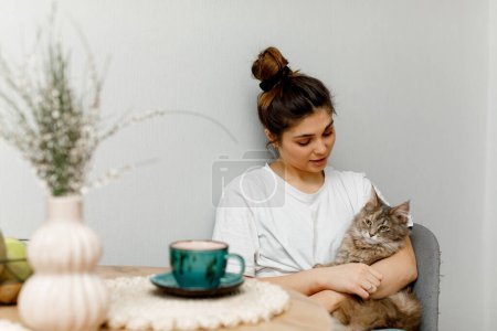 Photo for A brunette girl with hair styled in a bun is sitting in the kitchen with her cat and drinking tea. A girl in a white T-shirt is sitting at the table with a cup of green tea. - Royalty Free Image