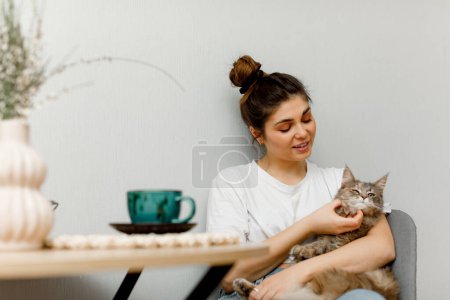Téléchargez les photos : Une fille brune coiffée dans un chignon est assise dans la cuisine avec son chat et boit du thé. Une fille en t-shirt blanc est assise à la table avec une tasse de thé vert. - en image libre de droit