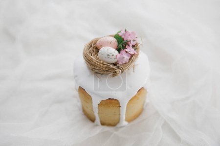 Téléchargez les photos : Gâteau de Pâques à glaçure blanche avec un nid d'oiseau décoratif avec des œufs et des fleurs sur une planche de bois et de la gaze blanche. Espace de copie. - en image libre de droit