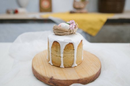 Téléchargez les photos : Gâteau de Pâques avec glaçure blanche avec un nid d'oiseau décoratif avec des œufs et des fleurs sur une planche de bois et gaze blanche. - en image libre de droit
