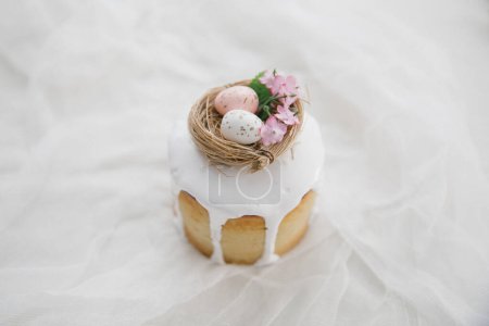 Foto de Pastel de Pascua con glaseado blanco con un nido de aves decorativo con huevos y flores en una tabla de madera y gasa blanca. Copiar espacio. - Imagen libre de derechos