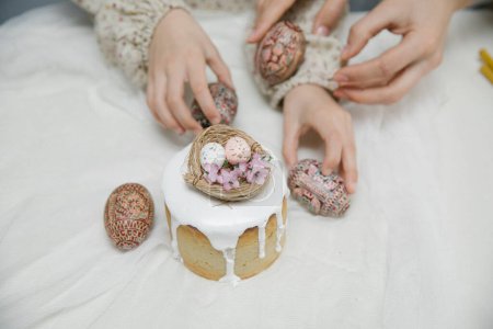 Téléchargez les photos : Gâteau de Pâques à glaçure blanche avec un nid d'oiseau décoratif avec des œufs et des fleurs. Œufs de Pâques en bois peint à la main dans les mains. - en image libre de droit
