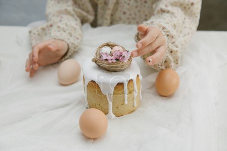 Photo for Easter cake with white glaze with a decorative bird's nest with eggs and flowers in child's hands. Child is holding Easter cake. - Royalty Free Image