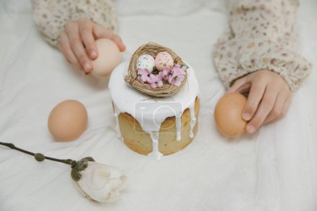 Téléchargez les photos : Gâteau de Pâques à glaçure blanche avec un nid d'oiseau décoratif avec des œufs et des fleurs dans les mains de l'enfant. Enfant tient gâteau de Pâques. - en image libre de droit