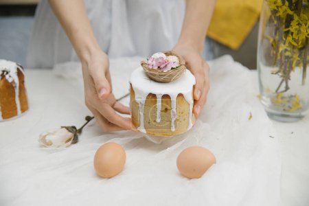 Foto de Pastel de Pascua con glaseado blanco con un nido de aves decorativo con huevos y flores en las manos. Mujer sosteniendo pastel de Pascua. - Imagen libre de derechos