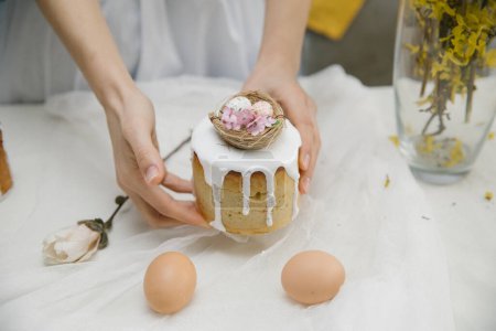 Foto de Pastel de Pascua con glaseado blanco con un nido de aves decorativo con huevos y flores en las manos. Mujer sosteniendo pastel de Pascua. - Imagen libre de derechos