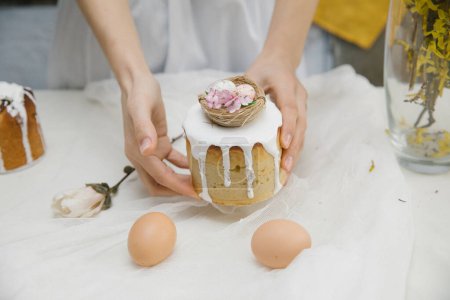 Téléchargez les photos : Gâteau de Pâques à glaçure blanche avec un nid d'oiseau décoratif avec des œufs et des fleurs dans les mains. Femme tenant gâteau de Pâques. - en image libre de droit