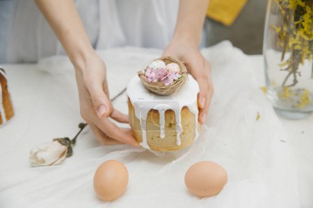 Téléchargez les photos : Gâteau de Pâques à glaçure blanche avec un nid d'oiseau décoratif avec des œufs et des fleurs dans les mains. Femme tenant gâteau de Pâques. - en image libre de droit