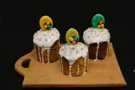 Téléchargez les photos : Gâteaux de Pâques avec glaçage blanc et décorations colorées sur une planche en bois sur fond noir. - en image libre de droit