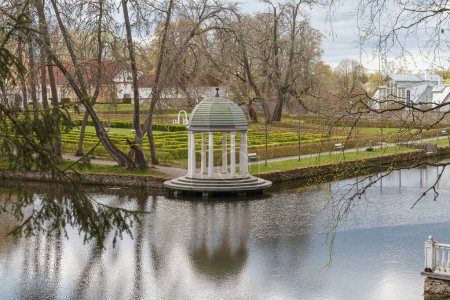 Classical white gazebo rotunda with round dome and columns reflected in the pond in a green park.