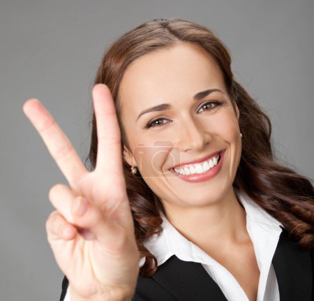 Téléchargez les photos : Joyeux sourire belle jeune femme d'affaires montrant deux doigts ou geste de victoire, sur fond gris - en image libre de droit