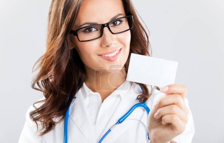 Téléchargez les photos : Portrait d'une jeune femme médecin souriante montrant une carte de visite vierge ou une invitation, sur fond gris - en image libre de droit