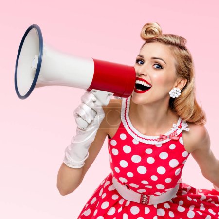 Photo for Portrait of woman holding megaphone, dressed in pin-up style red dress in polka dot and white gloves, over pink background. Caucasian blond model posing in retro fashion vintage studio shoot. - Royalty Free Image