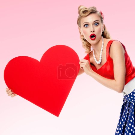 Photo for Woman holding heart symbol, dressed in pin-up style dress with polka dot, over pink background. Caucasian blond model posing in retro fashion and vintage concept studio shoot. - Royalty Free Image