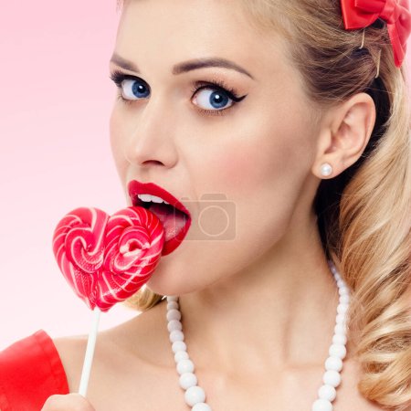 Téléchargez les photos : Portrait de femme mangeant sucette en forme de coeur vêtue d'une robe de style pinup à pois, sur fond rose. Modèle blond caucasien posant dans la mode rétro et vintage concept studio shoot. - en image libre de droit