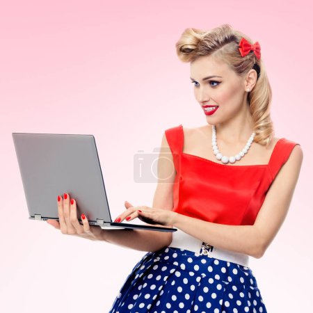 Photo for Portrait of beautiful woman holding laptop, dressed in pin-up style dress in polka dot, over pink background. Caucasian blond model posing in retro fashion and vintage concept studio shoot. - Royalty Free Image