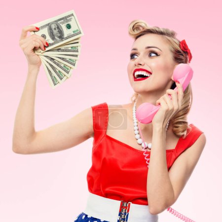 Photo for Happy woman with money, talking on phone, dressed in pin-up style dress in polka dot, over pink background. Caucasian blond model posing in retro fashion and vintage concept studio shoot. - Royalty Free Image
