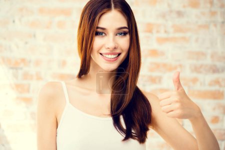 Foto de Mujer joven feliz en ropa blanca casual inteligente, mostrando los pulgares hacia arriba gesto, en el interior. Concepto publicitario. - Imagen libre de derechos