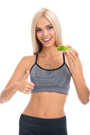 Photo for Woman in sportswear with vegetarian sandwich, showing thumb up, isolated over white background. Young sporty blond model at studio shot. Health, beauty and dieting concept. - Royalty Free Image