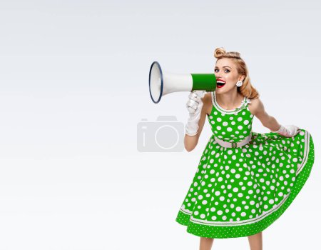 Porträt einer Frau mit Megafon, gekleidet in einem grünen Kleid im Pin-up-Stil mit Tupfen und weißen Handschuhen, auf grauem Hintergrund, mit leerem Kopierraum für Text oder Slogan. Kaukasische blonde Model posiert in Retro-Mode Vintage-Studio-Shooting.