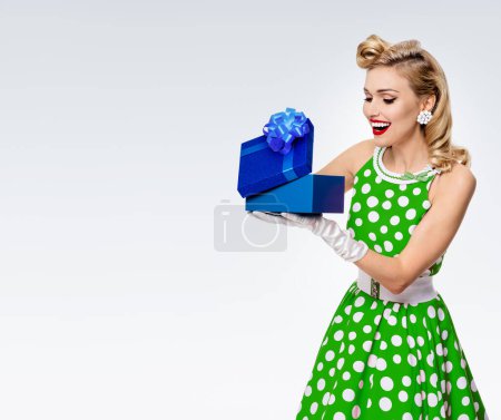 Portrait of beautiful young happy woman dressed in pin-up style green dress in polka dot and white gloves, on grey background, with blank copyspace area for text or slogan. Caucasian blond model posing in retro fashion studio shoot.