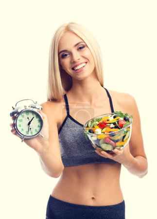 Photo for Woman in sportswear with plate of salad and alarm clock, isolated over yellow background. Young sporty blond model at studio shot. Healthy nutrition and time for dieting and fitness concept. - Royalty Free Image