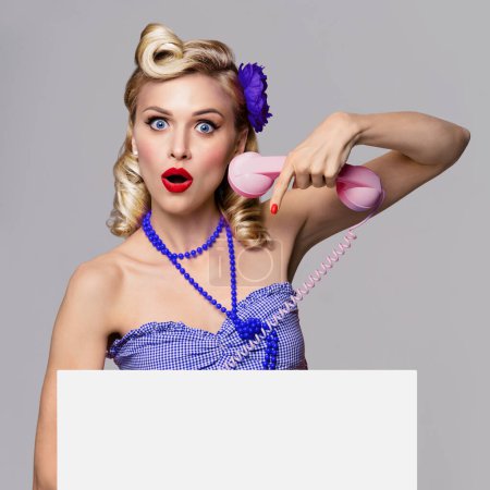 Photo for Portrait of beautiful young woman with phone and blank signboard, dressed in pin-up style. Caucasian blond model posing in retro fashion and vintage concept studio shoot, on grey background. - Royalty Free Image
