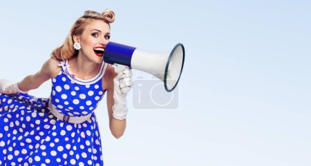 Portrait de femme tenant un mégaphone, vêtue d'une robe de style pin-up à pois et de gants blancs, avec espace pour slogan ou message publicitaire, sur fond bleu. Modèle blond caucasien posant dans la mode rétro vintage shoot.