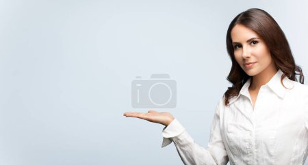 Happy smiling young cheerful businesswoman, showing something or blank copyspace area for slogan or text message, on grey background, with blank copyspace area for slogan or text. Success in business concept studio shot. 