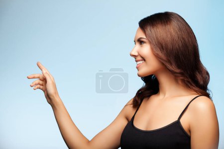 Photo for Young happy smiling brunette woman in black casual clothing, showing something or empty copyspace area for slogan or advertising text message, on bright blue background - Royalty Free Image