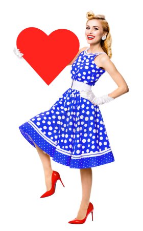 Photo for Full body of happy woman holding heart symbol, dressed in pin-up style blue dress with polka dot, isolated over white background. Caucasian blond model in retro fashion and vintage concept studio shoot. - Royalty Free Image