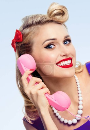 Foto de Retrato de la joven rubia sonriente feliz con teléfono, en estilo pin-up, sobre fondo azul. Modelo caucásico posando en retro moda y vintage concepto de rodaje de estudio. - Imagen libre de derechos