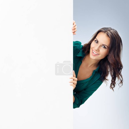Glücklich lächelnde junge Frau in grüner, selbstbewusster Kleidung, die vor grauem Hintergrund ein leeres Schild mit einer leeren Fläche für irgendeinen Text oder Slogan zeigt. Erfolg im Geschäfts- und Werbekonzept.