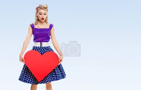 Photo for Full body of woman holding heart symbol, dressed in pin-up style dress with polka dot, with copyspace area for slogan or advertising text message, on blue background. Blond model in retro fashion and vintage concept. - Royalty Free Image