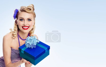 Portrait of beautiful young happy smiling woman in pin-up style clothing, with copyspace area for slogan or advertising text message, on blue background. Blond model in retro fashion and vintage concept.